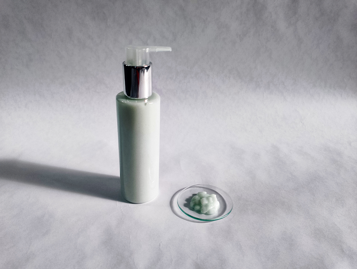 Telové mlieko v bielej fľaške s pumpičkou na bielom pozadí. Pred fľaškou je akrylová podložka s kvapkou telového mlieka.
